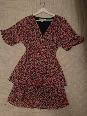 Blommig kortklänning med volang - Söt kortklänning med blommigt mönster i rosa och svart. Klänningen har v-ringning, volangdetaljer och korta ärmar. Perfekt för sommarens alla tillfällen. Tillverkad i ett lätt och luftigt material.