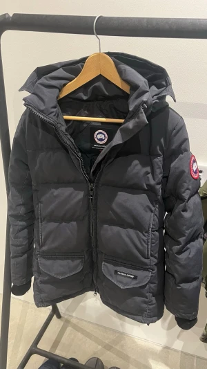 Svart pufferjacka från Canada Goose - Säljer en svart pufferjacka från Canada Goose med huva och dragkedja framtill. Jackan har två stora fickor med lock och den klassiska Canada Goose-loggan på ärmen. Perfekt för kalla vinterdagar och riktigt snygg streetstil.