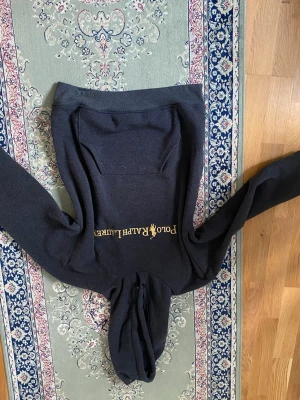 Svart hoodie från Polo Ralph Lauren - Säljer en svart hoodie från Polo Ralph Lauren med klassisk gul logga och text på bröstet. Hoodien har huva med dragsko och en stor magficka framtill. Perfekt för en avslappnad och stilren look.