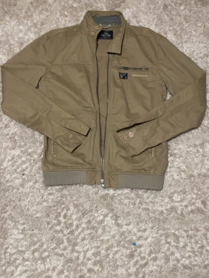 Vintage Limited edition Beige bomberjacka från Superdry - Snygg beige bomberjacka från Superdry med dragkedja framtill, ribbade muddar och två fickor med knappstängning. Jackan har en klassisk bomberpassform och är tillverkad i ett slitstarkt tyg. Limited Edition Tokyo-detalj på insidan. Står XL men passar M-L