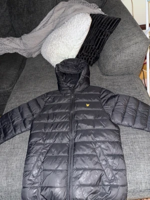 Svart dunjacka från Lyle & Scott - Snygg svart dunjacka från Lyle & Scott med gul logga på bröstet. Jackan har huva, dragkedja framtill och två sidofickor. Perfekt för kalla vinterdagar och har en klassisk quiltad design som håller värmen bra.