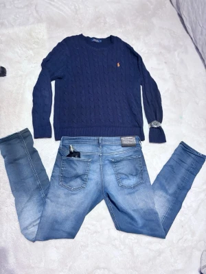 Mörkblå kabelstickad tröja Polo Ralph Lauren - Mörkblå kabelstickad tröja från Polo Ralph Lauren med rund halsringning och den klassiska broderade loggan i orange på bröstet. Tröjan har långa ärmar och ribbade muddar vid ärmslut och nederkant. Perfekt för en stilren och avslappnad look.