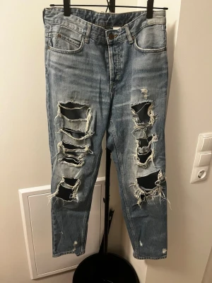 Ljusblå ripped jeans med stora hål - Säljer ett par ljusblå jeans med kraftiga slitningar och stora hål framtill. Jeansen har klassisk femficksmodell, hög midja och raka ben. Perfekta för en edgy streetstyle-look. Materialet är denim i bomull. Märke syns ej. Höger bakficka har ett större hål efter att ”trådarna” gick sönder.