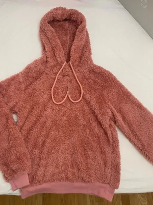 Rosa fluffig hoodie med huva - Supermjuk och fluffig hoodie i en härlig rosa färg. Hoodien har en stor huva med dragsko och breda muddar vid ärmslut och nederkant. Perfekt för dig som gillar mysiga och trendiga plagg. Materialet är teddy/fleece-liknande för extra komfort.