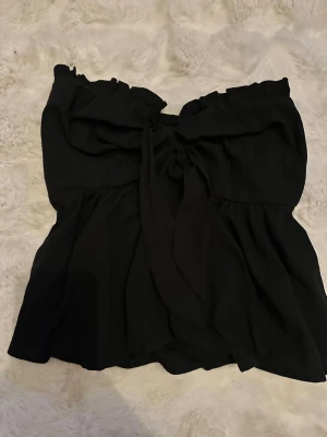 Svart axelbandslös tubtopp med knyt - Snygg svart axelbandslös tubtopp med volang och knyt framtill. Toppen har en luftig passform och är perfekt för varma dagar. Enkel att matcha med både jeans och kjol för en trendig look.