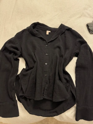 Svart skjorta med struktur - Säljer en svart skjorta med lång ärm och knappar framtill. Skjortan har en avslappnad passform, klassisk krage och är gjord i ett mjukt, strukturerat tyg som ger en cool look. Perfekt att styla lager på lager eller bära som den är.