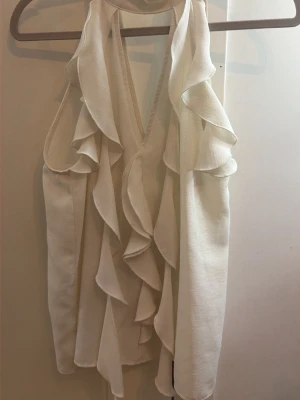 Vit volangtopp med v-ringning - Supersnygg vit topp med djup v-ringning och stora volangdetaljer framtill. Ärmlös modell som ger en fräsch och trendig look, perfekt att styla med jeans eller kjol. Tunt och mjukt material som känns skönt mot huden.