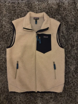 Patagonia fleeceväst - Snygg beige fleeceväst från Patagonia med dragkedja framtill, två sidofickor och en bröstficka i mörkblått tyg. Västen har hög krage och detaljer i blått. Perfekt för lager-på-lager och outdoor-stil.