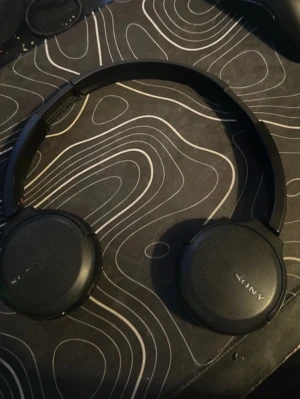 Svarta on-ear hörlurar från Sony - Snygga svarta on-ear hörlurar från Sony med rund design och justerbart huvudband. Hörlurarna har mjuka öronkuddar och en stilren look i plast och konstläder. Perfekta för musik, gaming eller videosamtal.