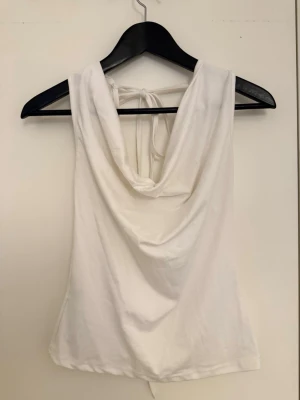 Vit linnetopp med snörning i ryggen - Säljer en vit linnetopp med snygg draperad urringning och snörning i ryggen. Toppen är ärmlös och har en mjuk, följsam passform som sitter bekvämt. Perfekt för dig som gillar minimalistisk och trendig stil.