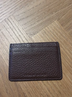 Brun skinnkorthållare tiger of Sweden! - Snygg och stilren korthållare från tiger of Sweden  i mörkbrunt skinn. Framsidan har präglad struktur och diskret Burberry-logga, medan baksidan är slät. Perfekt för kort och enkel att ha i fickan.💸