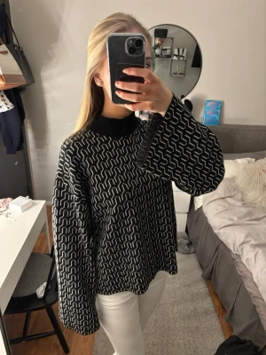 Svartvit stickad tröja med mönster - Snygg svart stickad tröja med vitt grafiskt mönster och rund hals. Tröjan har en avslappnad passform och vida ärmar, perfekt för en trendig och bekväm look. Materialet känns mjukt och passar bra till både jeans och kjol.