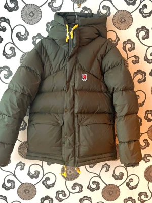 Grön dunjacka från Fjällräven down lite - Mörkgrön dunjacka från Fjällräven med stor huva och gula dragskor. Jackan har två stora framfickor med lock, tryckknappar och dragkedja. Fjällrävens klassiska rävlogga sitter på bröstet. Perfekt för kalla vinterdagar.