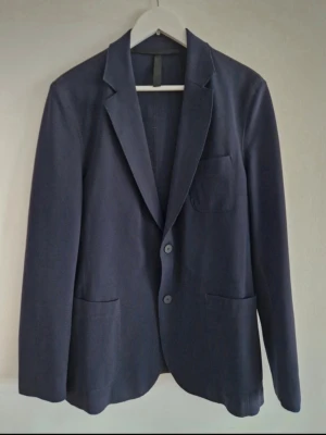 J.Lindeberg mörkblå overshirt - Stilren mörkblå overshirt med två knappar framtill, tre utanpåliggande fickor och klassisk krage. Overshirten har en rak passform och är tillverkad i ett mjukt tyg som ger en avslappnad men ändå dressad look. Perfekt att bära över en t-shirt eller skjorta.