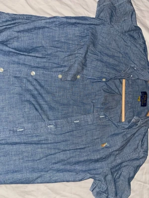 Blå kortärmad skjorta Polo Ralph Lauren - Snygg blå kortärmad skjorta från Polo Ralph Lauren i mjukt bomullstyg. Klassisk button-down-krage och broderad logga på bröstet. Perfekt till jeans eller shorts för en avslappnad men stilren look. Som förra skjortan exakt samma storlek säljer för att jag behöver pengarna just nu. Knappt använd endast någon enstaka gång mycket bra skick. 