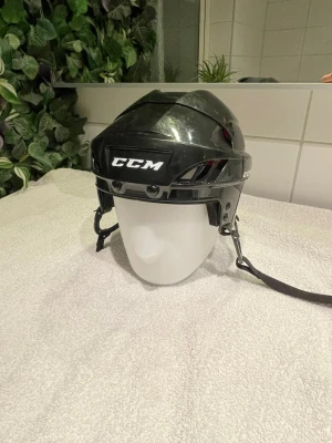 Hockeyhjälm från CCM stl. Medium - Säljer en svart hockeyhjälm från CCM med hakrem och ventilationshål. Hjälmen har röda detaljer samt logotyp framtill. Nytt skick då den knappast har blivit använd.