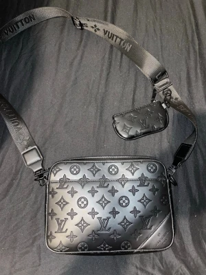 Svart axelväska Louis Vuitton - Snygg svart axelväska från Louis Vuitton i präglat läder med klassiskt monogrammönster. Väskan har justerbar axelrem med logotyp och en liten avtagbar miniväska. Perfekt för dig som vill ha en stilren och lyxig accessoar.