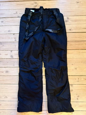 Svarta täckbyxor med hängslen - Säljer ett par svarta täckbyxor med justerbara hängslen. Använda för skidåkning. Säljs billigt pga smutsiga och lite slitna och noppriga (se bilder)