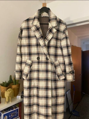 Rutig lång rock från Zara - Snygg lång rock från Zara i beige, svart och brunt rutigt mönster. Rocken har dubbelknäppning, bred krage och två stora fickor framtill. Perfekt för dig som gillar klassisk stil med en modern twist.
