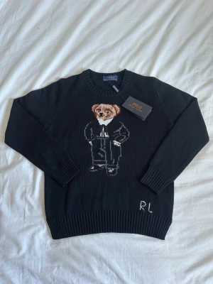 Svart stickad tröja Polo Ralph Lauren - Svart stickad tröja från Polo Ralph Lauren med ikonisk björn i kostym broderad på bröstet. Rund halsringning och ribbade muddar vid ärmslut och nederkant. RL-broderi nedtill. Perfekt för dig som gillar klassisk men lekfull stil.