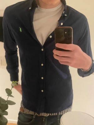 Mörkblå skjorta från Polo Ralph Lauren - Mörkblå skjorta från Polo Ralph Lauren i slim fit-modell. Skjortan har klassisk krage, knappar framtill och en liten grön broderad logga på bröstet. Perfekt för en stilren och avslappnad look.