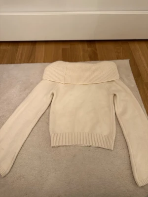 Off shoulder stickad tröja cremevit - Trendig off shoulder stickad tröja i cremevit färg med bred ribbad krage och långa ärmar. Tröjan har en croppad passform och ribbade muddar vid ärmslut och nederkant. Perfekt för en stilren och modern look.