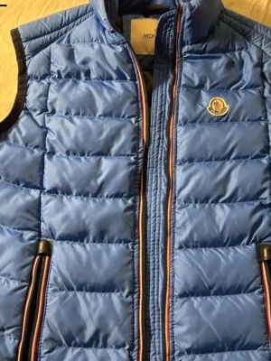 Blå dunväst från Moncler - Snygg blå dunväst från Moncler med quiltad design och klassisk logotyp på bröstet. Västen har svarta detaljer vid ärmöppningarna och dragkedjor med färgade kanter. Perfekt för lager-på-lager och streetstyle.