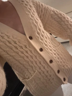 Beige kabelstickad kofta - Kabelstickad beige kofta med stora bruna knappar och två framfickor. Koftan har djup V-ringning och ribbade muddar. Perfekt för lager på lager och ger en mysig, trendig look. Materialet är mjukt och värmande.