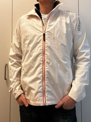 Vit vindjacka från Helly Hansen - Stilren vit vindjacka från Helly Hansen med hel dragkedja framtill och diskreta röda detaljer. Jackan har trycket 'SPEED PERFORMANCE' på ena ärmen och logga på axeln. Lätt material och hög krage med gul detalj på insidan. Längd: 70cm, P2P: 50cm