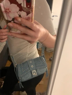 Ljusblå jeans axelväska med nitar - Trendig ljusblå 2000-tals axelväska i jeansmaterial med silverfärgade nitar och justerbar rem. Väskan har en cool spänndetalj framtill och dekorativa fransar på sidorna. Perfekt för dig som gillar en edgy och unik stil. Köpt för lite mer än 1000kr utomlands men kom aldrig till användning därav säljare jag billigt. Den har inga tecken på skada/fläckar. Pris kan diskuteras vid snabb affär!