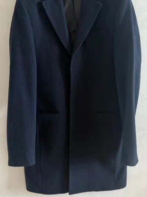 Mörkblå ullkappa från Hampton Republic - Säljer en Hampton Republic kappa i storlek 46. Kappan är i väldigt bra skick utan några skador. Den har tre knappar i framsidan med en ficka på högra bröstet. Inuti kappan finns det ytterligare två fickor på högra sidan samt en på vänstra sidan. Kappan kommer med en extra knapp ifall det skulle behövas. Längst ner på kappans baksida är den designad med en slits/öppning. Jag skickar gärna fler bilder och svarar på frågor om det skulle uppstå några. Priset går att diskuteras!