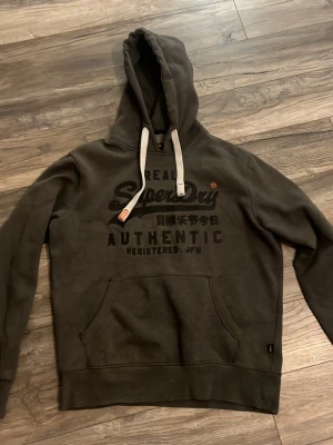 Mörkgrå Superdry hoodie med tryck - Säljer en mörkgrå hoodie från Superdry med stor logga och japansk text på bröstet. Hoodien har vita dragsnören, känguruficka och en klassisk huva. Perfekt för dig som gillar streetwear och vill ha en bekväm och snygg tröja.