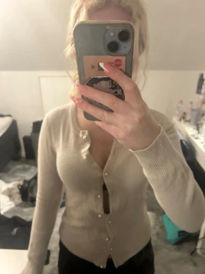 Beige kofta med pärlknappar - Snygg beige kofta med långa ärmar och ribbade muddar. Koftan har v-ringning och stängs med dekorativa pärlknappar framtill. Perfekt att styla till både jeans och kjol.