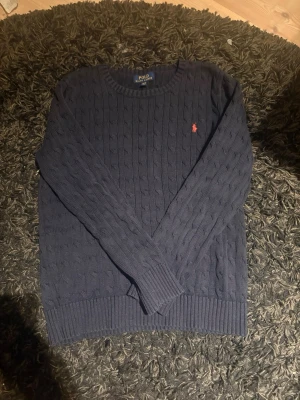 Mörkblå kabelstickad tröja Polo Ralph Lauren - Kabelstickad mörkblå tröja från Polo Ralph Lauren med rund halsringning och den klassiska röda logotypen broderad på bröstet. Tröjan har ribbade muddar vid ärmslut och nederkant, perfekt för en stilren och tidlös look.