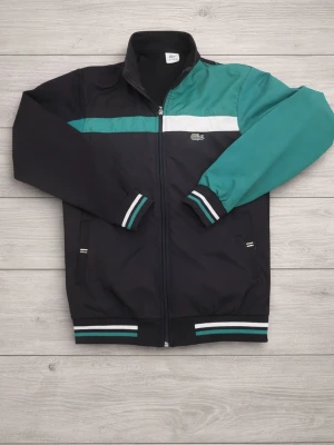 Lacoste bomberjacka svart/grön - Snygg bomberjacka från Lacoste i svart med gröna och vita detaljer. Jackan har dragkedja framtill, ribbade muddar med randigt mönster och en kontrasterande grön ärm. Klassisk Lacoste-logga på bröstet och stor logga på ryggen. Perfekt för en sportig streetwear-look.