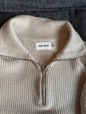 Beige stickad tröja med halvzip - Säljer en beige stickad tröja från EAST WEST med halvzip och hög krage. Tröjan har lång ärm och ribbstickad struktur som ger en clean och stilren look. Perfekt för lager på lager och enkel att matcha med olika outfits. Bara använd ett fåtal gånger! Skriv vid funderingar😁