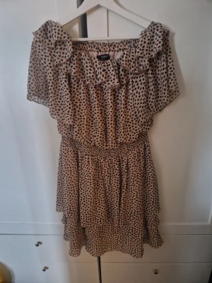 Leopardmönstrad klänning Sisters Point - Snygg kort klänning från Sisters Point i beige med svart leopardmönster. Offshoulder-modell med volang upptill och resår i midjan för en bekväm passform. Perfekt för dig som gillar trendiga detaljer och djurmönster.