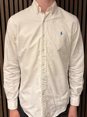 Vit skjorta från Polo Ralph Lauren - Klassisk vit skjorta från Polo Ralph Lauren med blå broderad logga på bröstet. Skjortan har button-down krage, långa ärmar och knäppning framtill. Modellen är custom fit och tillverkad i bomull, perfekt för en clean och stilren look.
