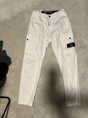 Vita cargobyxor från Stone Island - Snygga vita cargobyxor från Stone Island med klassisk patch på benet. Byxorna har raka ben, stora fickor med lock och knappstängning på sidorna samt en bekväm midja. Perfekta för en streetwear-look och tillverkade i ett slitstarkt bomullsmaterial. Jag ör 183 och byxorna passar mig bra 