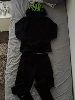 Syna world tracksuit - Säljer ett svart loungewear-set med hoodie, byxor och strumpor. Hoodien har grön graffiti-text på huvan och byxorna har matchande grön graffiti på baksidan. Setet är i mjukt material och har en avslappnad passform med dragsko i midjan. Perfekt för chill dagar hemma.