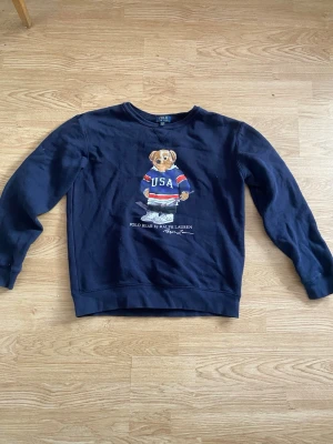 Mörkblå Polo Bear hoodie Ralph Lauren - Mörkblå hoodie från Ralph Lauren med den ikoniska Polo Bear-figuren på bröstet, klädd i en USA-tröja och hockeyutrustning. Rund halsringning och mjukt material som känns skönt mot huden. Perfekt för dig som gillar sportig och klassisk stil.