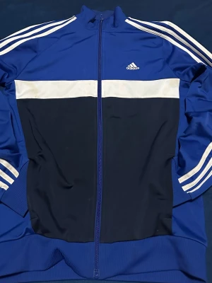 Adidas blå träningsjacka med dragkedja - Blå träningsjacka från Adidas med vita ränder på ärmarna och vit panel över bröstet. Jackan har hel dragkedja, hög krage och ribbade muddar. Tillverkad i funktionsmaterial med AEROREADY-tryck på ryggen. Perfekt för sport eller chill.