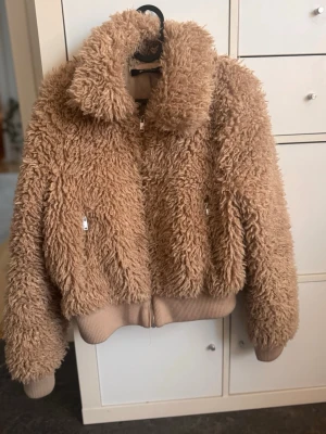 Beige teddyjacka med dragkedja - Mysig beige teddyjacka med fluffig struktur och hög krage. Jackan har dragkedja framtill, två fickor med dragkedja och ribbade muddar vid ärmar och nederkant. Perfekt för dig som vill ha en varm och trendig look.