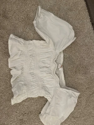 Vit croppad topp från Gina Tricot - Söt vit croppad topp från Gina Tricot med smockdetaljer och korta puffärmar. Toppen har en elastisk passform och vågig kant upptill, perfekt för en trendig och avslappnad look.