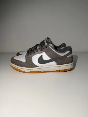 Nike Dunk Low grå/vit sneakers - Nike Dunk Low sneakers i grått och vitt med mörkgrå Swoosh och gummisula. Ovandelen är i mocka och textil, med klassisk perforerad tåbox och grå skosnören. Perfekt för dig som gillar stilrena och tidlösa sneakers. Som man kan se bilden så har den några defekter.