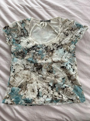 Blommig mesh topp med knyt i nacken - Snygg blommig mesh topp i vitt, blått och beige med transparent material och knyt i nacken. Toppen har korta ärmar och en rundad urringning med spetsdetaljer. Perfekt för dig som gillar unika mönster och lätta material.