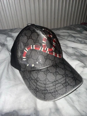 Gucci keps - Snygg svart keps från Gucci med klassiskt GG-mönster och en röd, vit och svart orm broderad på framsidan. Kepsen har mesh på sidorna och justerbar rem bak med gröna och röda detaljer. Perfekt för dig som vill sticka ut med en lyxig accessoar.