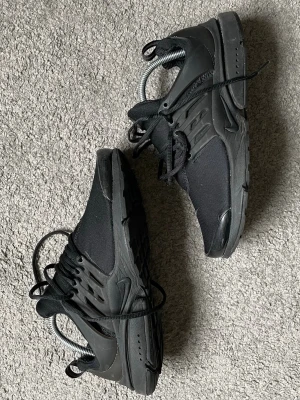 Nike Presto svart sneakers - Säljer ett par helsvarta Nike Presto sneakers med mesh-överdel och plastdetaljer vid snörningen. Skorna har en låg profil och en flexibel sula som ger grym komfort. Perfekt för dig som gillar en clean och sportig look. Lite sönder, se bild 