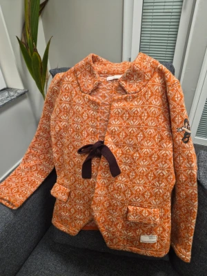 Orange mönstrad kofta Odd Molly storlek 2 S - Säljer en orange och vit stickad kofta från Odd Molly med grafiskt mönster, två fickor fram och brun knytning. Koftan har en broderad detalj på ärmen och en liten etikett vid ena fickan. Perfekt för dig som gillar färg och unika detaljer.