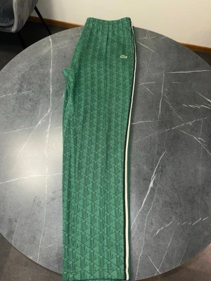 Gröna mjukisbyxor från Lacoste - Säljer ett par gröna mjukisbyxor från Lacoste med diskret mönster och vita sidoränder. Byxorna har elastisk midja och det klassiska Lacoste-märket på höften. Perfekta för en chill och sportig look.